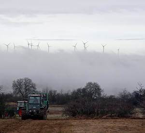 Traktor fährt auf landwirtschaftlicher Fläche. Im Hintergrund Nebel und Windenergieanlagen.